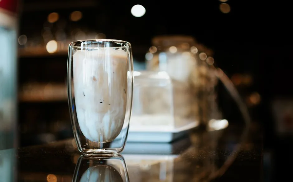 A glass of iced coffee on a reflective counter in a cozy café setting with warm lighting and bokeh effect.