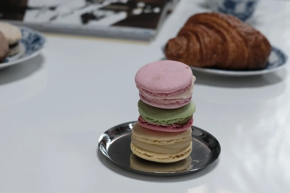A stack of colorful macarons on a plate with a croissant in the background, perfect for a French pastry theme.