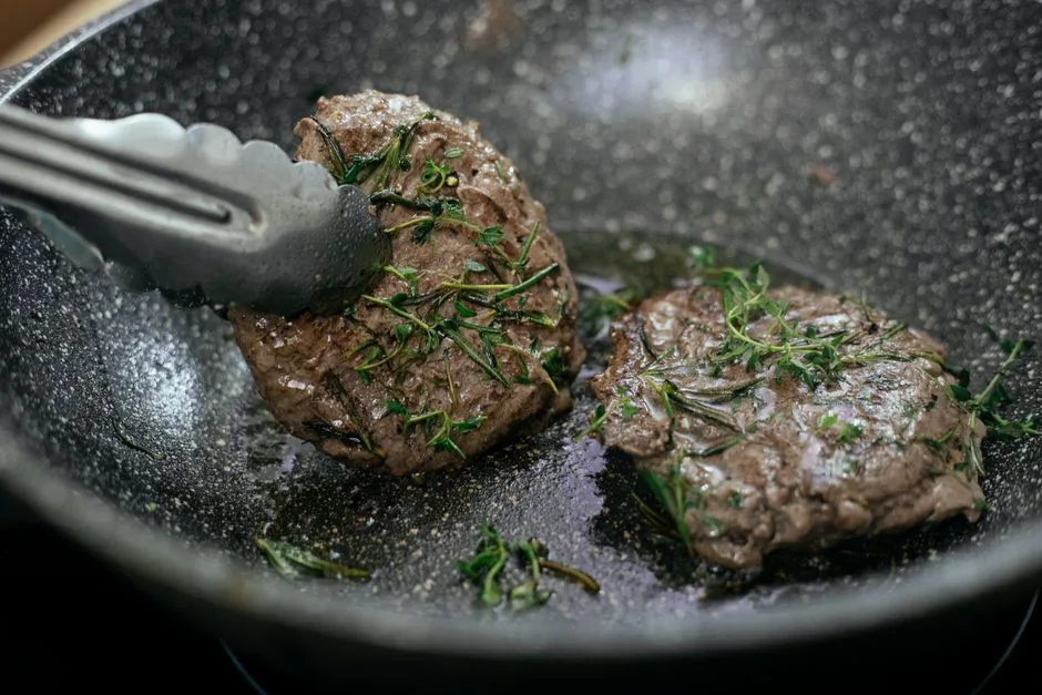 Juicy cutlets topped with aromatic rosemary frying in hot pan with metal tongs during cooking process in kitchen while preparing for lunch