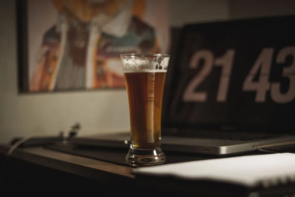 A glass of beer on a desk with a laptop showing time in the background offers a cozy evening workspace vibe.