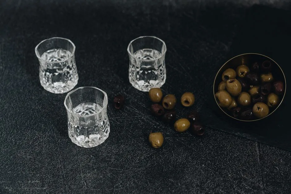 Top view of crystal glasses filled with vodka and bowl of olives on a dark textured surface.