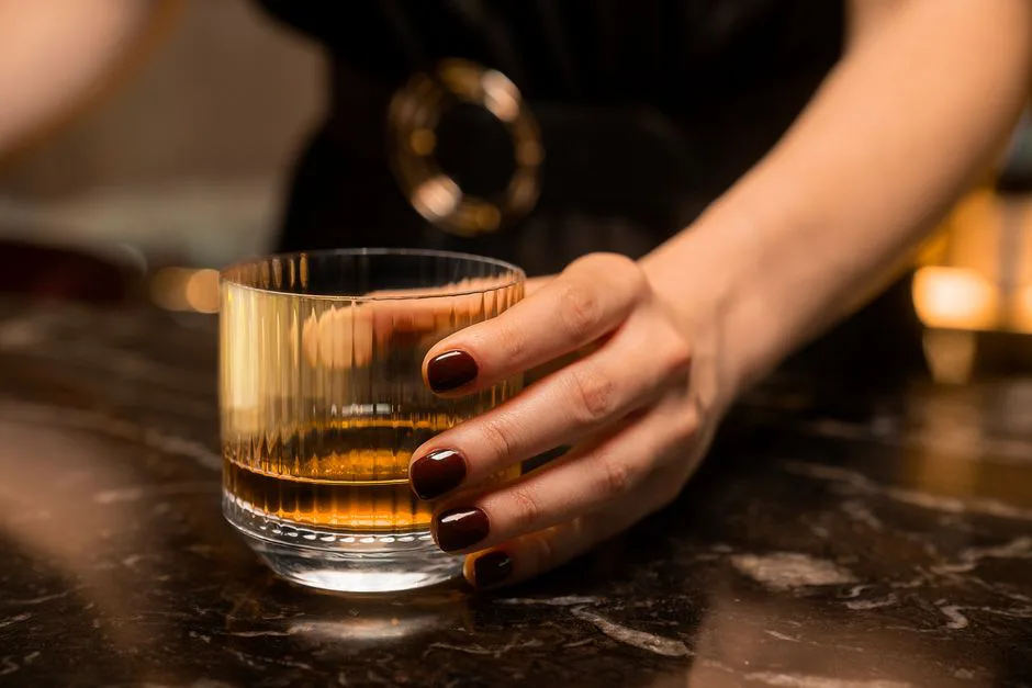Close-up of a woman's hand holding a glass of whiskey on a marble table, with warm lighting.