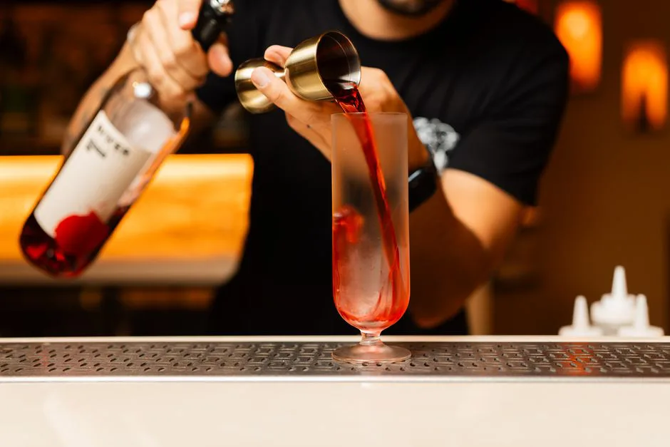 Close-up of a bartender pouring a vibrant red cocktail into a glass at a bar counter.