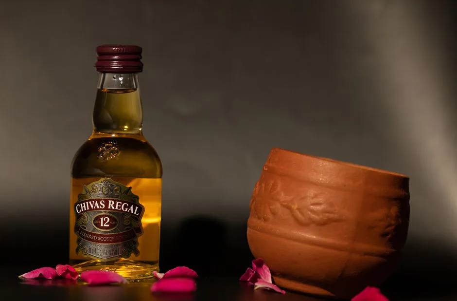 Chivas Regal 12 year whisky with clay cup and rose petals on dark background.