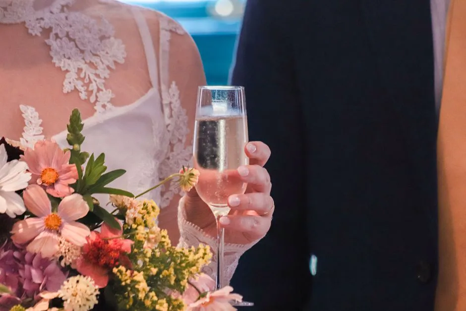 Bride in lace holding a floral bouquet and champagne glass at wedding ceremony.