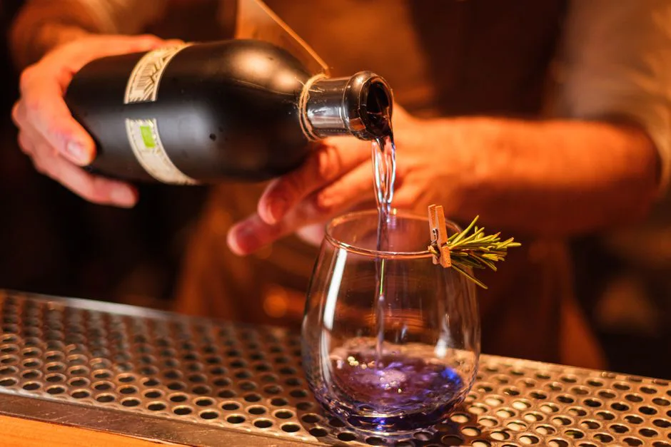 A bartender pours a drink into a glass garnished with rosemary at a dimly-lit bar.