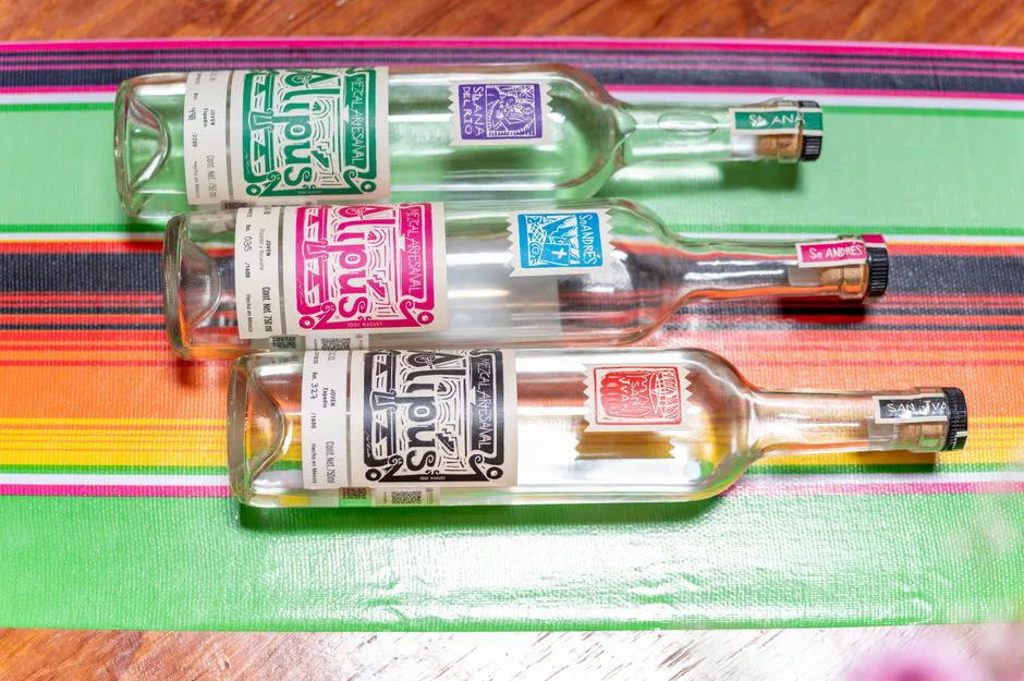 Three artisanal mezcal bottles on a vibrant Mexican blanket, showcasing traditional Mexican drink culture.