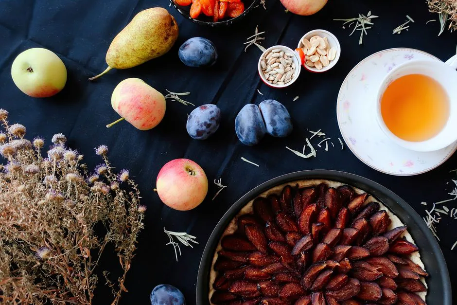 Flat lay of colorful fruits, plum tart, and herbal tea on a dark table setting.