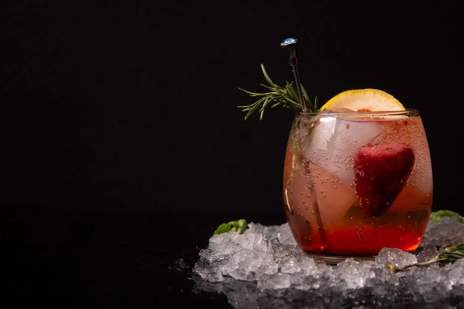 Chilled cocktail with lemon and strawberry garnish on ice against black background.