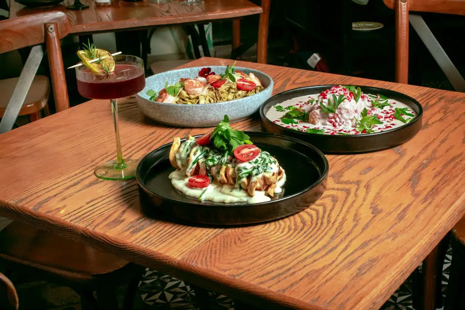 Three gourmet dishes with a cocktail served on a wooden table, showcasing culinary art in a cozy restaurant setting.