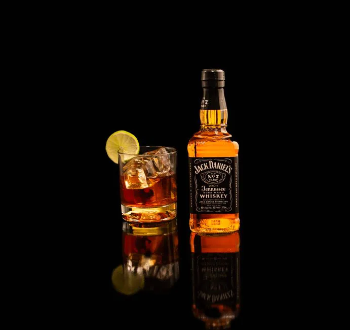 Jack Daniel's bottle and whiskey glass with lime on reflective black surface.