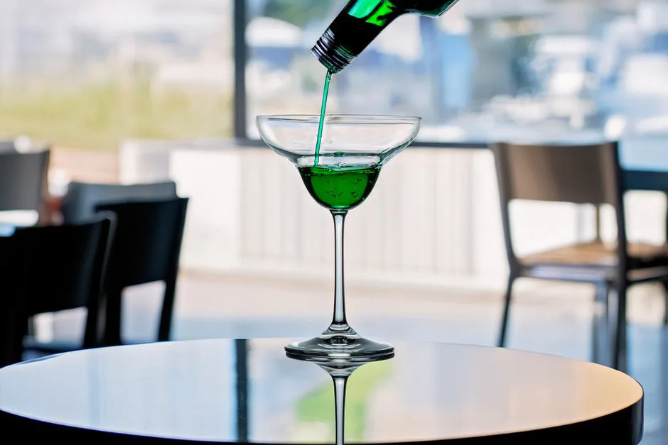 A vibrant green cocktail being poured into a glass indoors with blurred background.